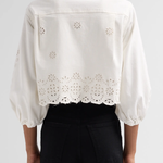 A person shown from the back wearing a white eyelet cropped blouse with puff sleeves and black pants against a light gray studio background.