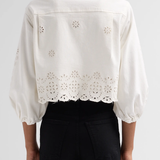 A person shown from the back wearing a white eyelet cropped blouse with puff sleeves and black pants against a light gray studio background.