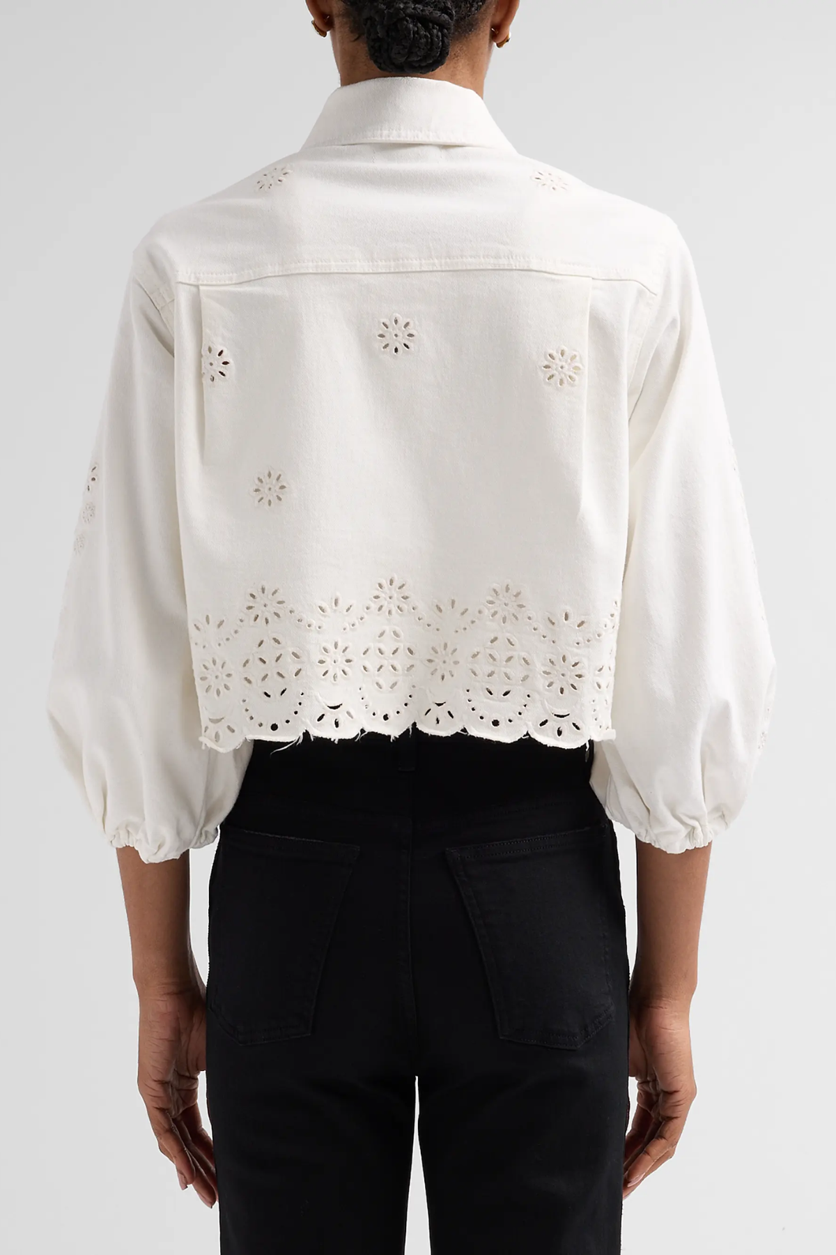 A person shown from the back wearing a white eyelet cropped blouse with puff sleeves and black pants against a light gray studio background.