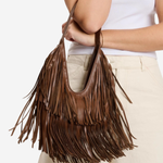 A brown leather fringe shoulder bag held by a model wearing a white ribbed tank top and beige pants, shown against a plain white background.