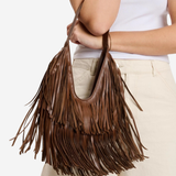 A brown leather fringe shoulder bag held by a model wearing a white ribbed tank top and beige pants, shown against a plain white background.