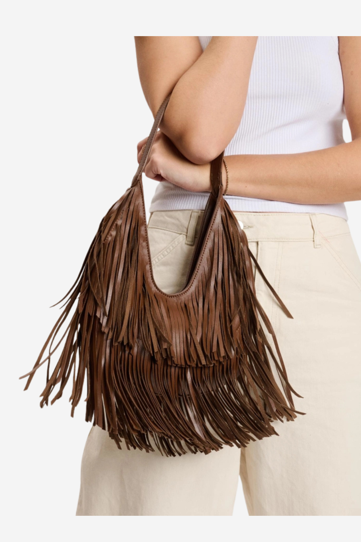 A brown leather fringe shoulder bag held by a model wearing a white ribbed tank top and beige pants, shown against a plain white background.