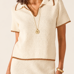 A woman wearing a cream-colored textured knit short-sleeve top with brown trim, paired with a matching knit skirt. She is accessorized with gold necklaces and a bracelet against a plain light background.