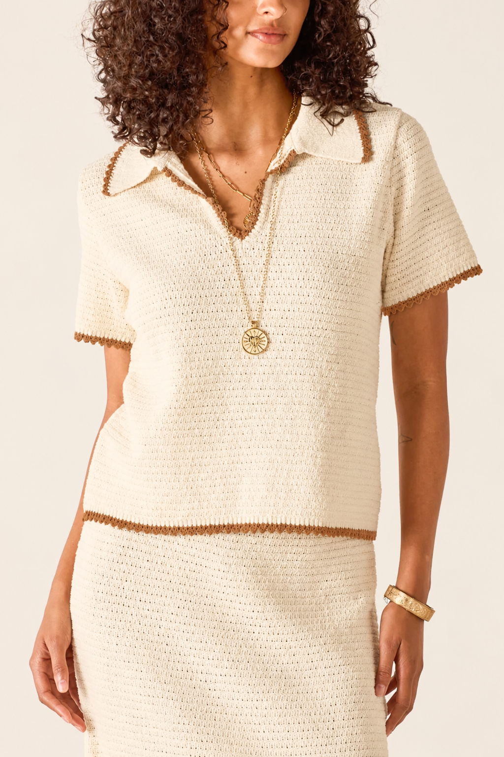 A woman wearing a cream-colored textured knit short-sleeve top with brown trim, paired with a matching knit skirt. She is accessorized with gold necklaces and a bracelet against a plain light background.