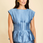 A woman modeling a light blue textured satin-like top with cap sleeves and a gathered peplum waist, paired with matching blue pants, against a solid beige background.