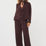 A model wearing a dark brown double-breasted blazer and matching wide-leg trousers, styled with a dark patterned neck scarf and white shoes, against a plain light gray studio background.