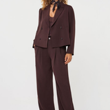 A model wearing a dark brown double-breasted blazer and matching wide-leg trousers, styled with a dark patterned neck scarf and white shoes, against a plain light gray studio background.