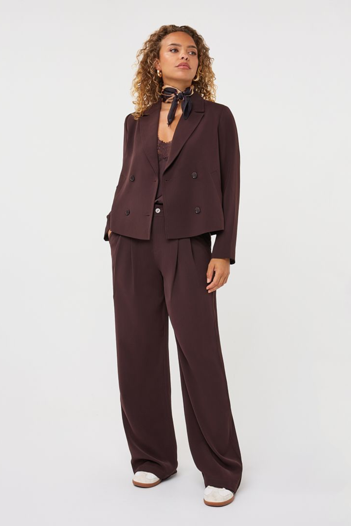 A model wearing a dark brown double-breasted blazer and matching wide-leg trousers, styled with a dark patterned neck scarf and white shoes, against a plain light gray studio background.