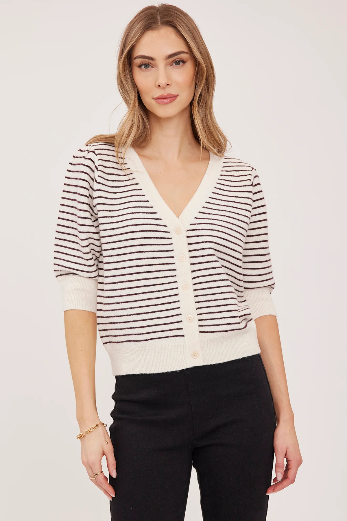 A woman modeling a short-sleeve knit cardigan with thin dark brown horizontal stripes on an off-white base, featuring a V-neck and button-front closure, paired with black pants against a plain light background.