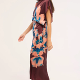 A woman modeling a short-sleeve midi dress with a dark burgundy base and a large abstract floral print in peach, pink, and blue. She is wearing beige heeled mule sandals against a plain light background.