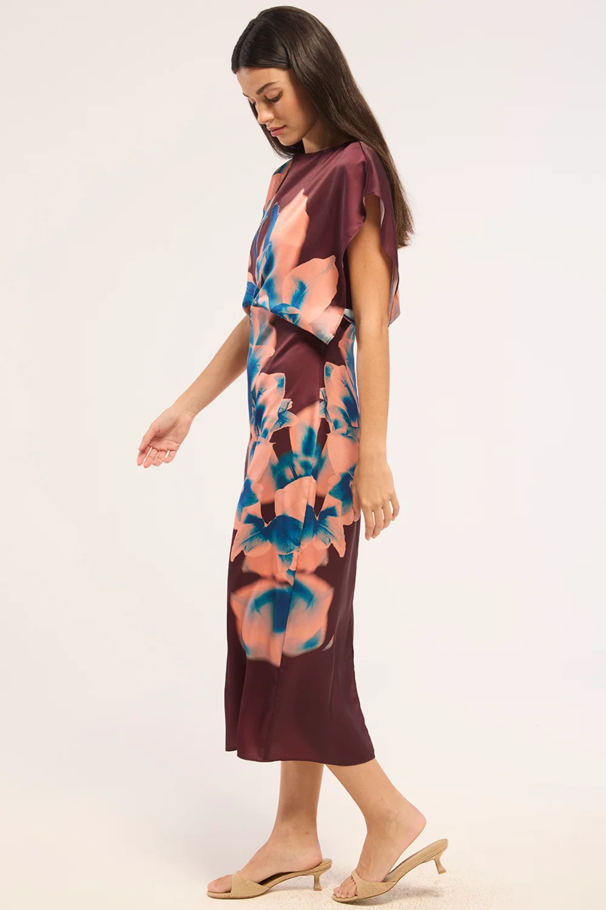 A woman modeling a short-sleeve midi dress with a dark burgundy base and a large abstract floral print in peach, pink, and blue. She is wearing beige heeled mule sandals against a plain light background.
