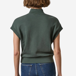 A woman viewed from the back wearing a dark olive green short-sleeve mock-neck knit top and blue jeans against a plain light background.