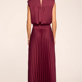 A woman shown from the back wearing a sleeveless pleated maxi dress in a deep burgundy or wine color, paired with beige high-heeled sandals, against a plain light beige studio background.