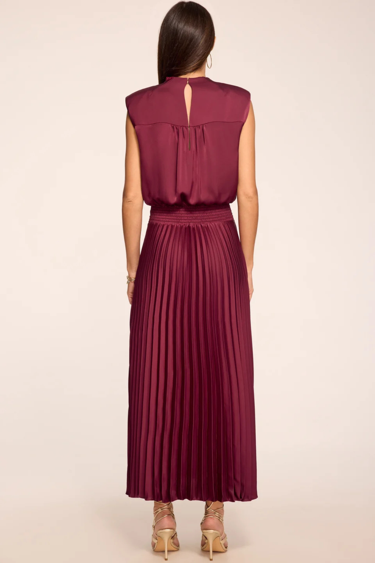 A woman shown from the back wearing a sleeveless pleated maxi dress in a deep burgundy or wine color, paired with beige high-heeled sandals, against a plain light beige studio background.