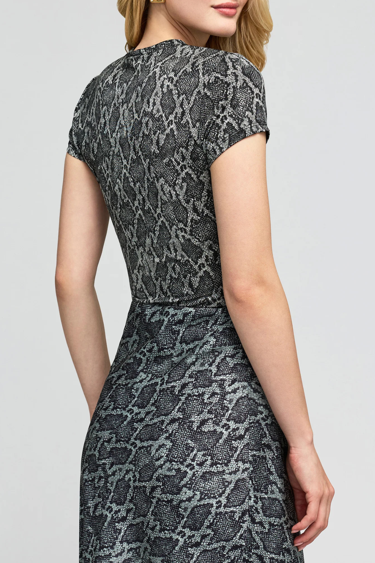 A woman wearing a short-sleeve fit-and-flare dress with a gray and black snakeskin pattern, shown from a rear three-quarter angle against a light gray studio background.
