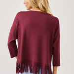 A woman shown from the back wearing a maroon long-sleeve top with fringe trim at the hem, paired with light blue jeans, against a plain light gray background.
