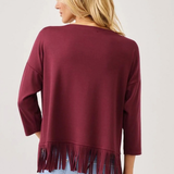 A woman shown from the back wearing a maroon long-sleeve top with fringe trim at the hem, paired with light blue jeans, against a plain light gray background.
