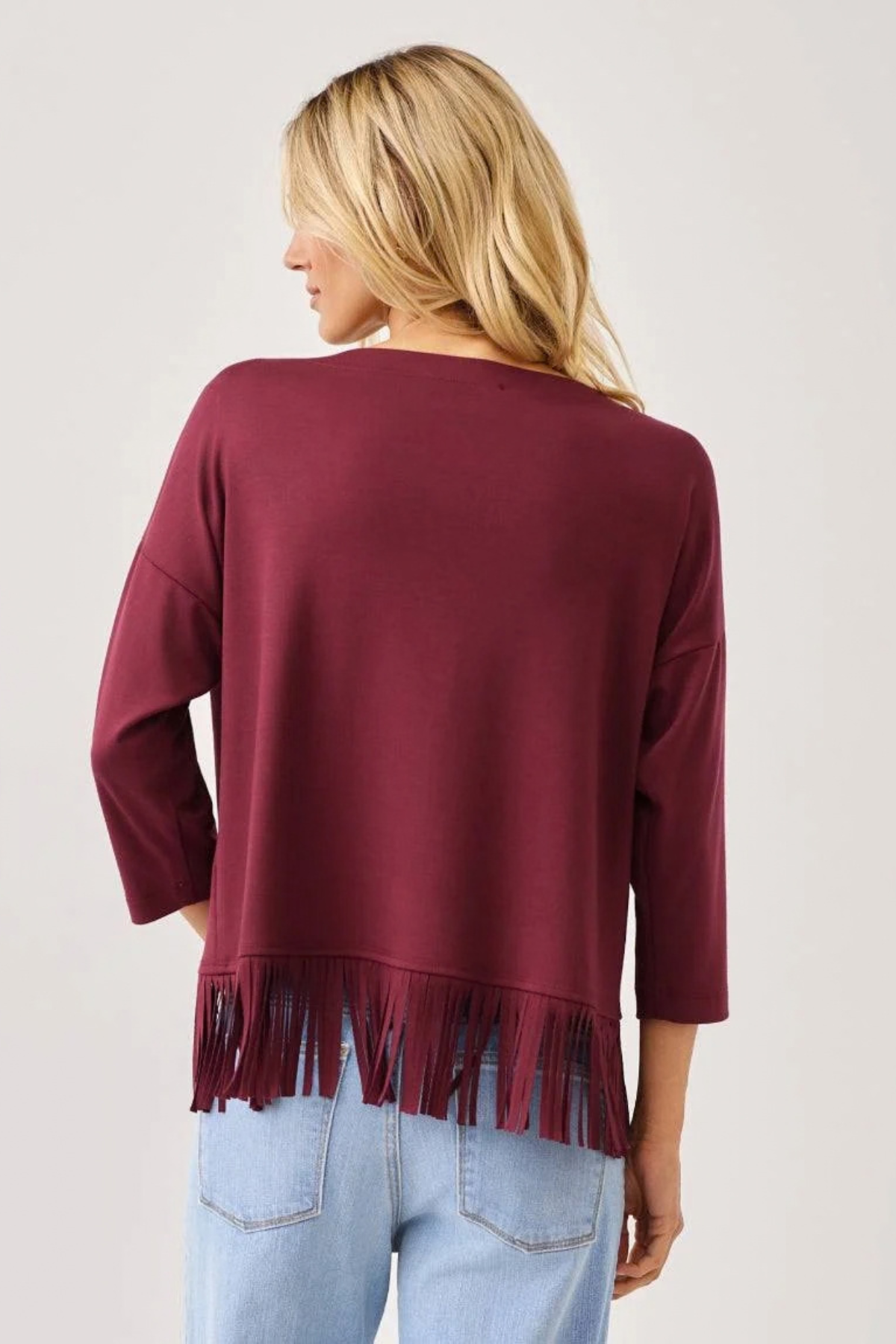 A woman shown from the back wearing a maroon long-sleeve top with fringe trim at the hem, paired with light blue jeans, against a plain light gray background.