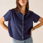 A woman modeling a short-sleeve navy blue button-up shirt with white contrast stitching and light-wash blue jeans, standing against a plain light beige background.