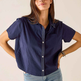 A woman modeling a short-sleeve navy blue button-up shirt with white contrast stitching and light-wash blue jeans, standing against a plain light beige background.
