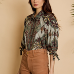 A woman modeling a long-sleeve button-up blouse with a detailed bird and floral pattern in olive green, brown, and beige, paired with brown trousers, set against a warm beige background with subtle plant leaves at the corners.