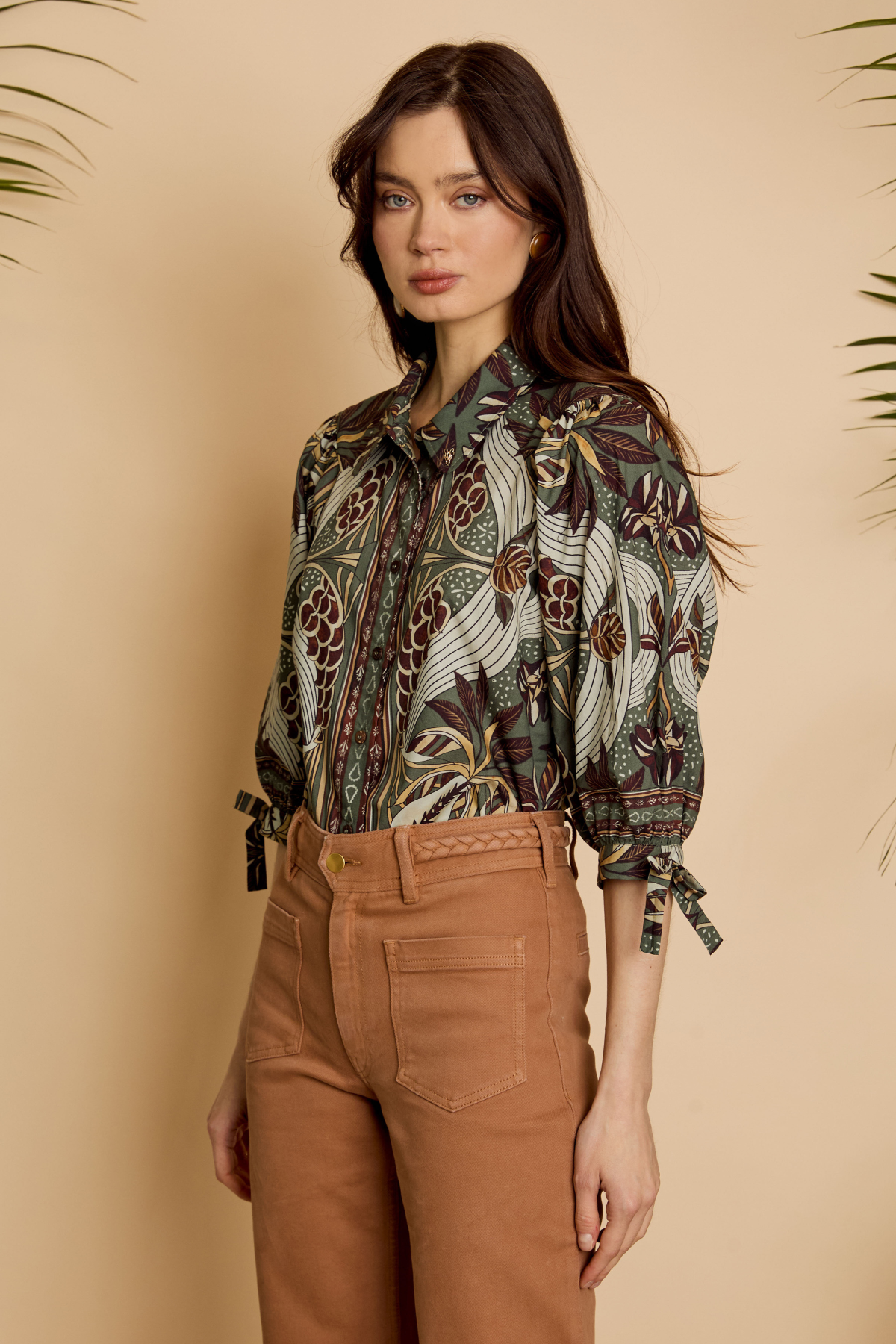 A woman modeling a long-sleeve button-up blouse with a detailed bird and floral pattern in olive green, brown, and beige, paired with brown trousers, set against a warm beige background with subtle plant leaves at the corners.