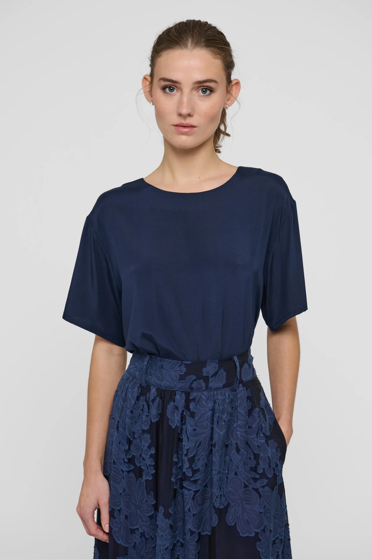 A woman modeling a navy blue short-sleeve top with a matching navy floral lace skirt against a light gray studio background.