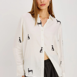 A woman modeling a white button-up shirt with black dog silhouettes printed all over it, paired with black pants, against a plain light gray studio background.