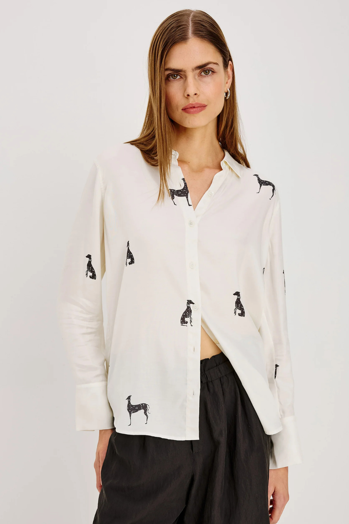 A woman modeling a white button-up shirt with black dog silhouettes printed all over it, paired with black pants, against a plain light gray studio background.