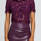 A woman wearing a short-sleeve purple lace button-up shirt with puff sleeves and a shiny purple faux-leather pencil skirt, shown from the nose down against a light gray studio background.
