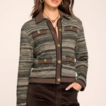A woman models a cropped, long-sleeve tweed jacket with a textured green, brown, and gray pattern, featuring brown lapels, button details, and flap pockets. She is also wearing dark brown pants against a solid light beige studio background.