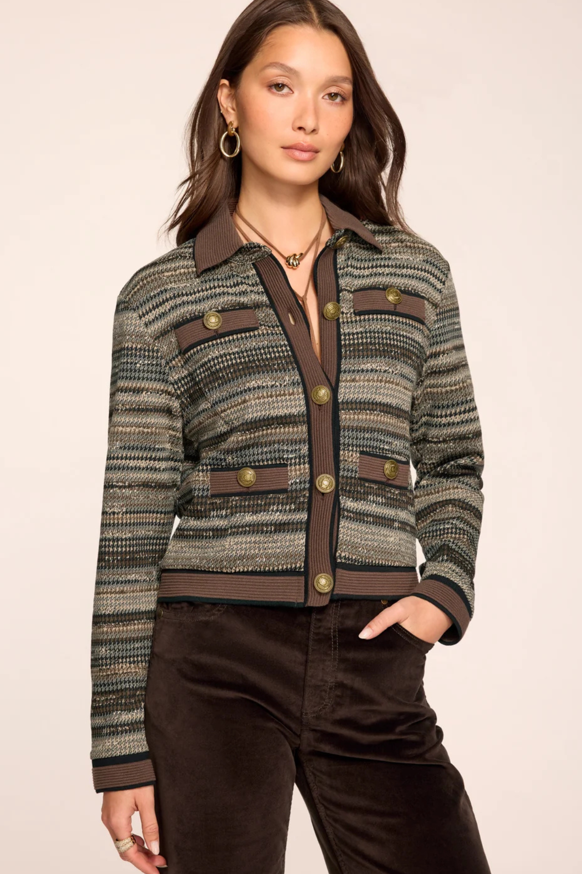A woman models a cropped, long-sleeve tweed jacket with a textured green, brown, and gray pattern, featuring brown lapels, button details, and flap pockets. She is also wearing dark brown pants against a solid light beige studio background.