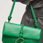 Green handbag with a gold clasp held by a person wearing gray pants on a light gray background