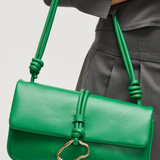 Green handbag with a gold clasp held by a person wearing gray pants on a light gray background