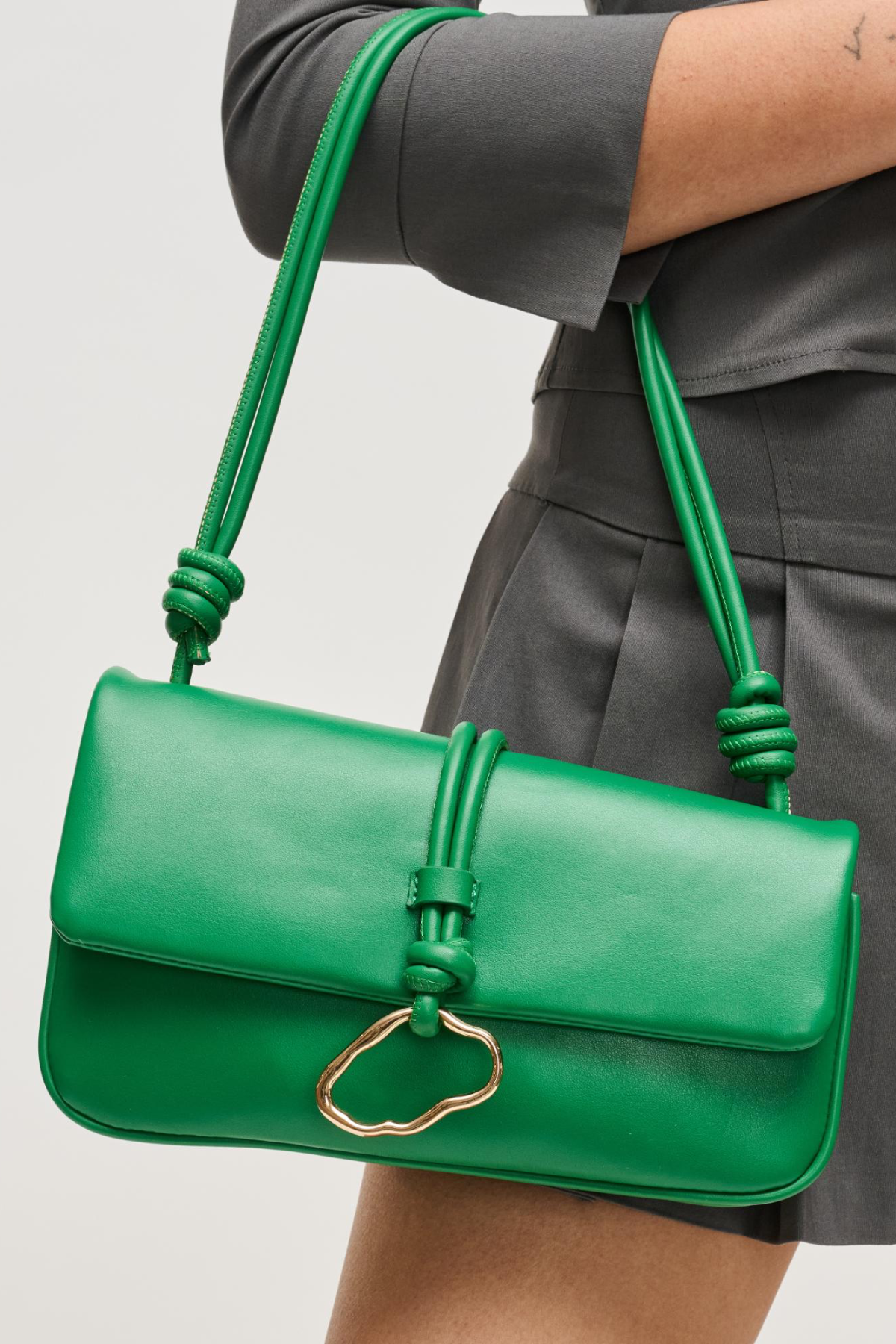 Green handbag with a gold clasp held by a person wearing gray pants on a light gray background
