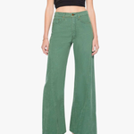 A model wearing a pair of sage green wide-leg denim pants with a black mesh crop top and gray heeled sandals, photographed against a plain white studio background.