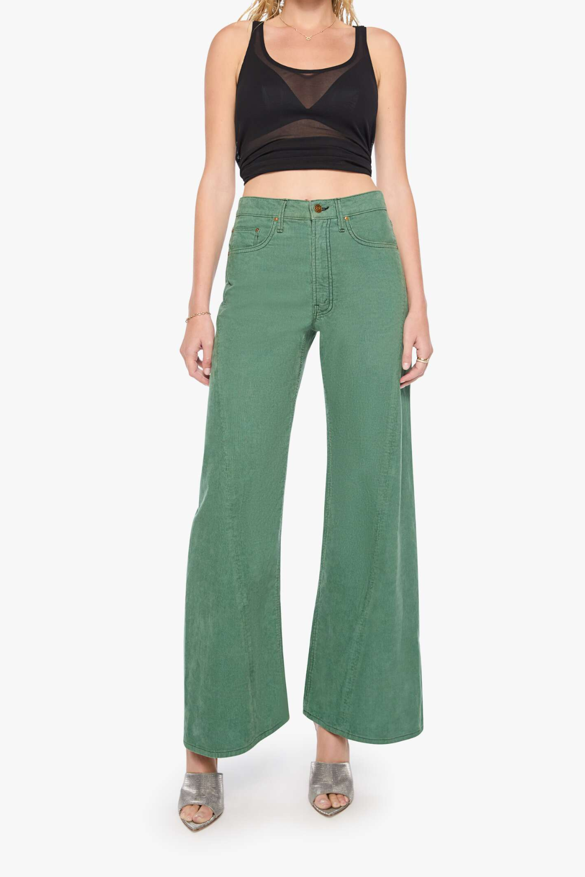 A model wearing a pair of sage green wide-leg denim pants with a black mesh crop top and gray heeled sandals, photographed against a plain white studio background.