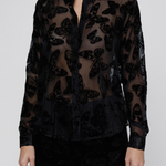 A model wearing a sheer black long-sleeve button-up blouse with a black butterfly pattern, paired with black pants, against a light gray studio background.