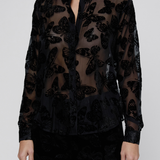 A model wearing a sheer black long-sleeve button-up blouse with a black butterfly pattern, paired with black pants, against a light gray studio background.
