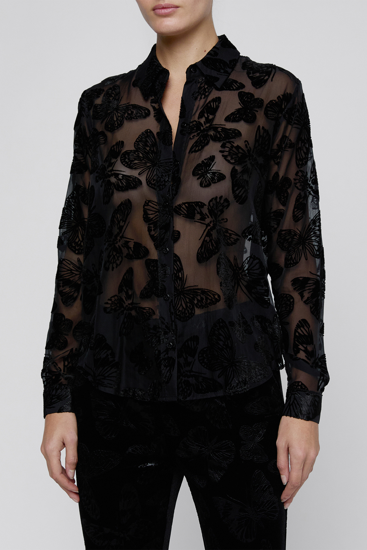 A model wearing a sheer black long-sleeve button-up blouse with a black butterfly pattern, paired with black pants, against a light gray studio background.