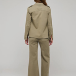 A woman viewed from the back wearing a matching khaki military-style uniform set consisting of a long-sleeve button-up shirt and wide-leg trousers, paired with black flat sandals, against a light gray studio background.