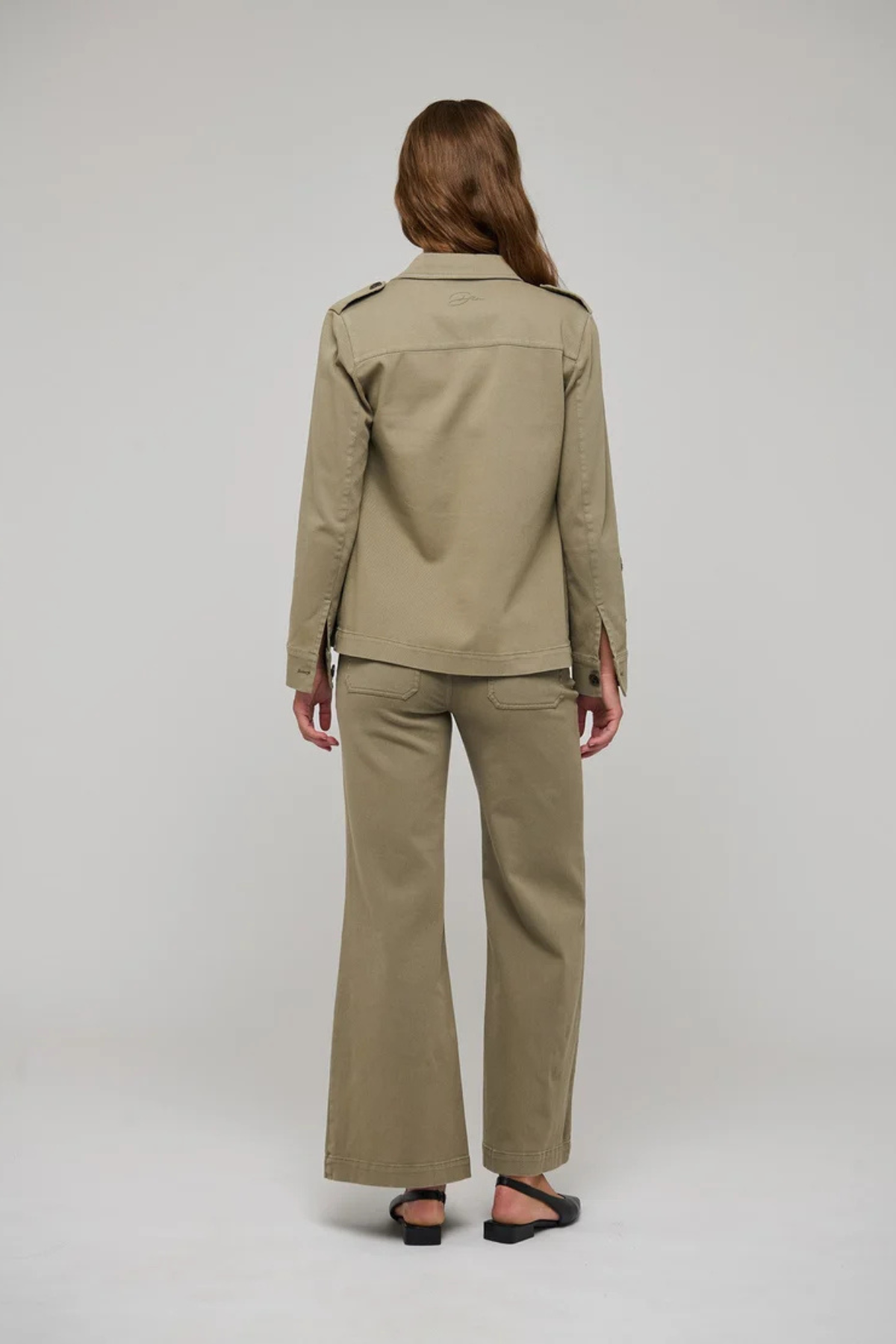 A woman viewed from the back wearing a matching khaki military-style uniform set consisting of a long-sleeve button-up shirt and wide-leg trousers, paired with black flat sandals, against a light gray studio background.