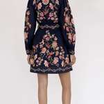A woman shown from the back wearing a navy blue long-sleeve mini dress with a colorful floral and bird print, paired with black strappy sandals against a plain white studio background.