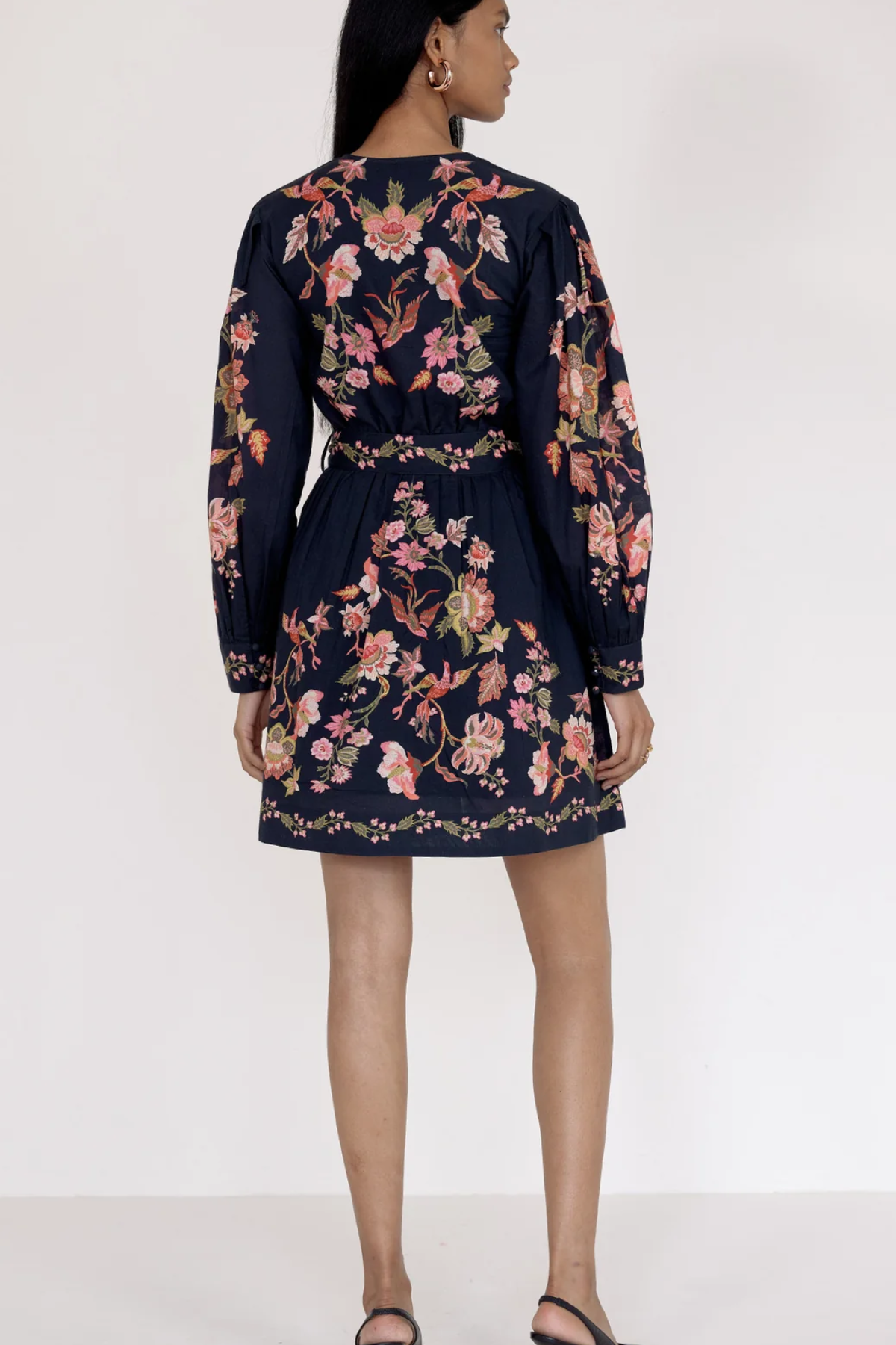 A woman shown from the back wearing a navy blue long-sleeve mini dress with a colorful floral and bird print, paired with black strappy sandals against a plain white studio background.