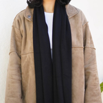 A woman wearing a beige suede-like jacket with a black scarf and a white top, standing against a plain off-white wall.