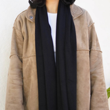 A woman wearing a beige suede-like jacket with a black scarf and a white top, standing against a plain off-white wall.