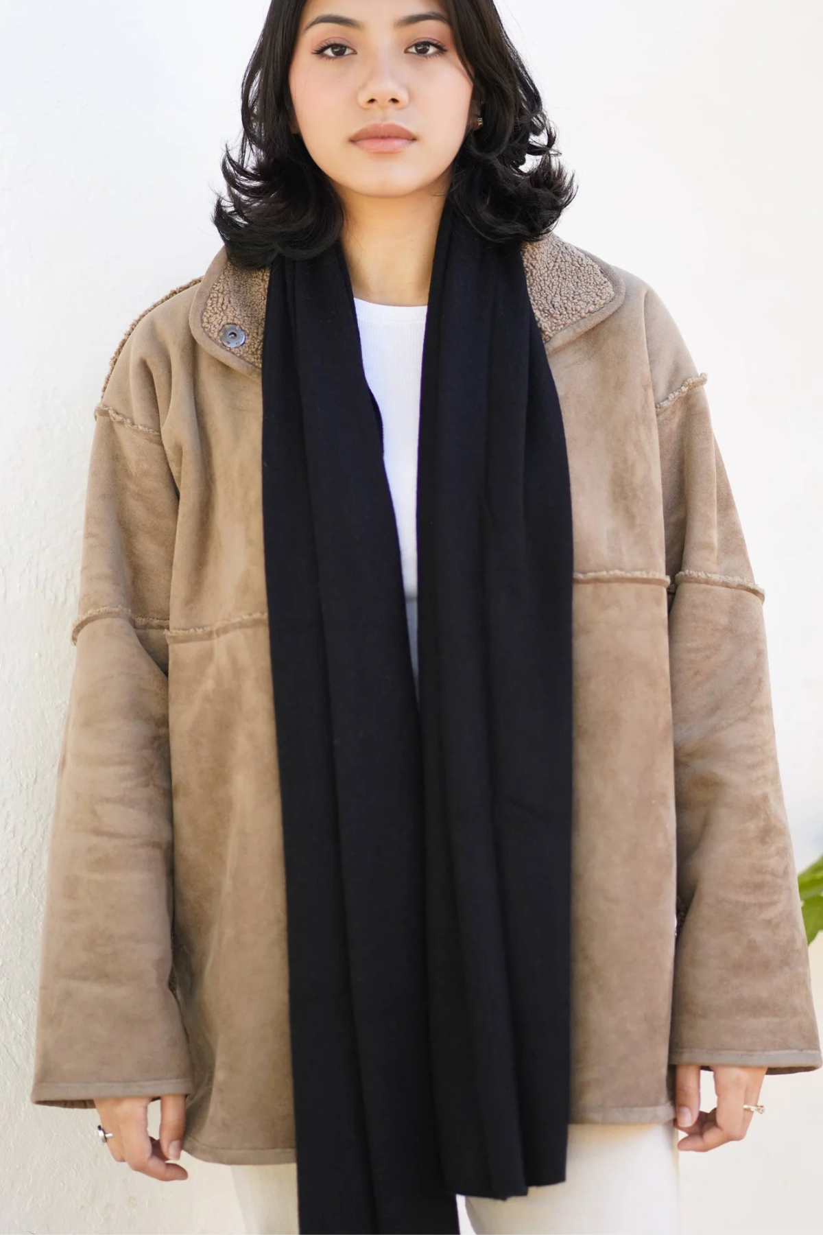 A woman wearing a beige suede-like jacket with a black scarf and a white top, standing against a plain off-white wall.