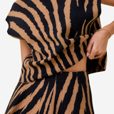 A close-up of a model wearing a matching two-piece knit set with a bold black and tan tiger-stripe pattern. The set includes a high-neck, short-sleeved top and a matching flared skirt.