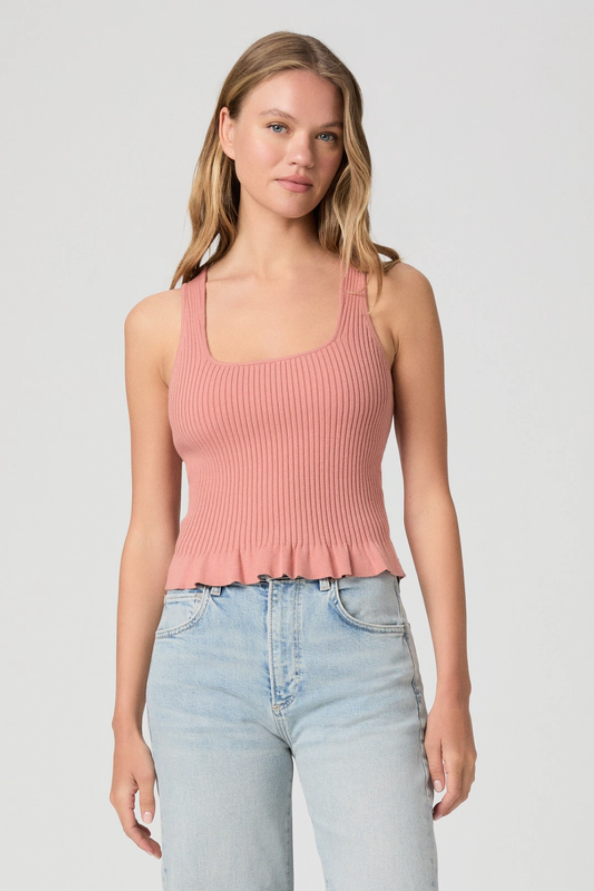 Woman wearing a pink ribbed tank top and light blue jeans on a plain background