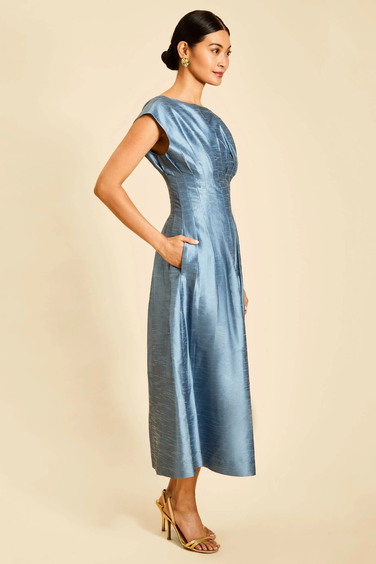 A woman modeling a light blue satin midi dress with cap sleeves and side pockets, standing against a solid beige background.